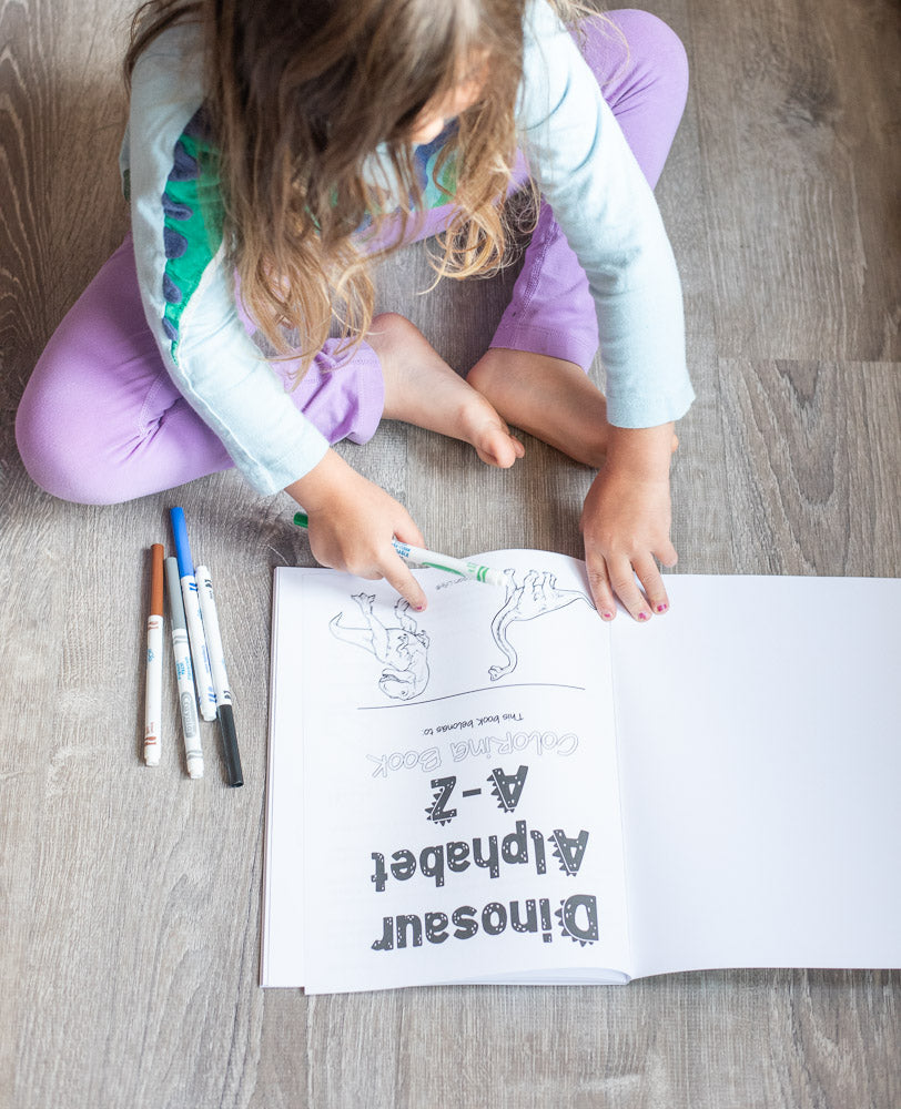 A preschooler wearing a dino shirt with a Dinosaur Alphabet A-Z coloring book on the floor